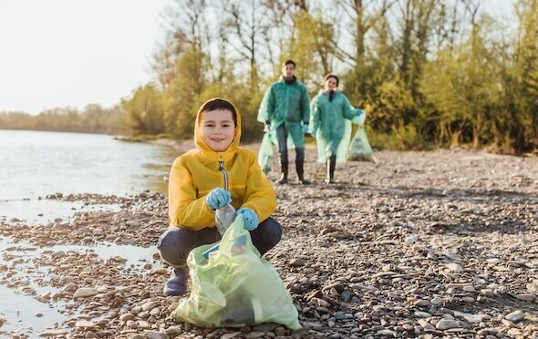 Image showing young boy in a yellow coat, crouching on a shallow river bank with a plastic bag of litter. His parents are walking behind also carrying litter bags.