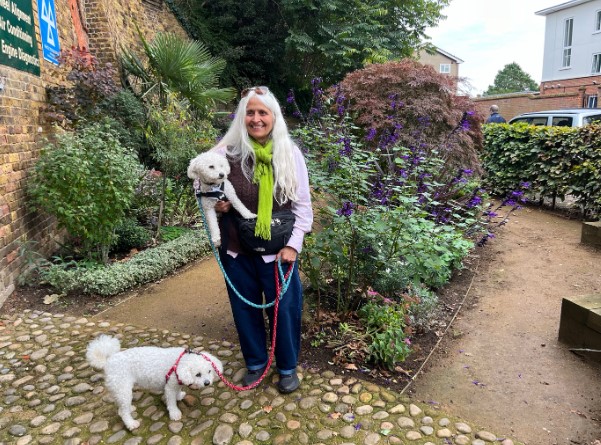 Image of Bharti Hindocha smiling and standing in the gardens holding one white poodle under her arm with another on a lead by her feet.