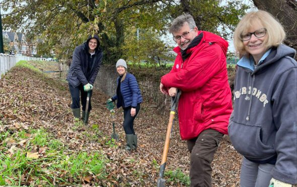 Four people smiling at the camera and holding spades, standing in the slope in front of the Ha ha.