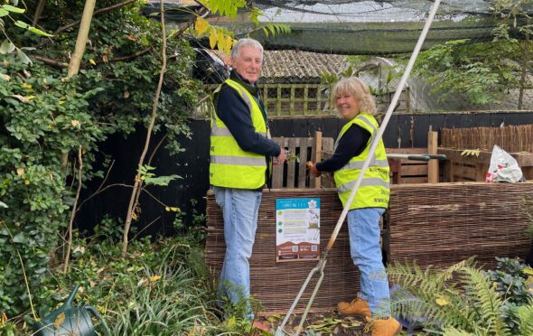 Image shows two gardeners, a man and a woman, in yellow hi-viz jackets, blue jeans and black shirts standing either side of an A4 Composting 1-2-3 poster. The poster is fixed on to the front a composting bin. The man is on the left and the woman is on the right. Both are dropping garden waste into the left hand bin of a dual compost bin system. There are shrubs and plants around their feet and a tree to the left of the image