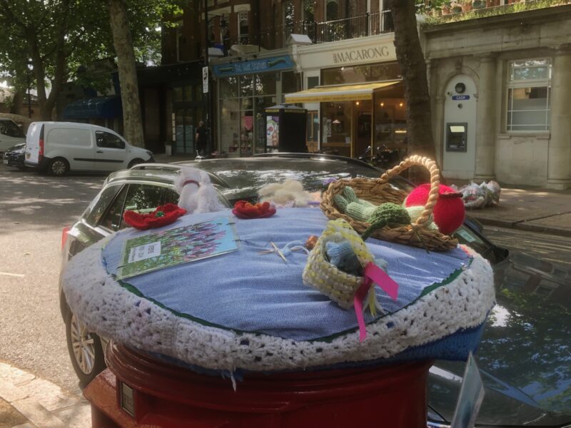 Image shows a knitted topper on a red postbox. The topper includes a knitted basket of fruit, dog, flowers and basket or wool. There is a row of shops in the background and a parked car directly behind the post box.
