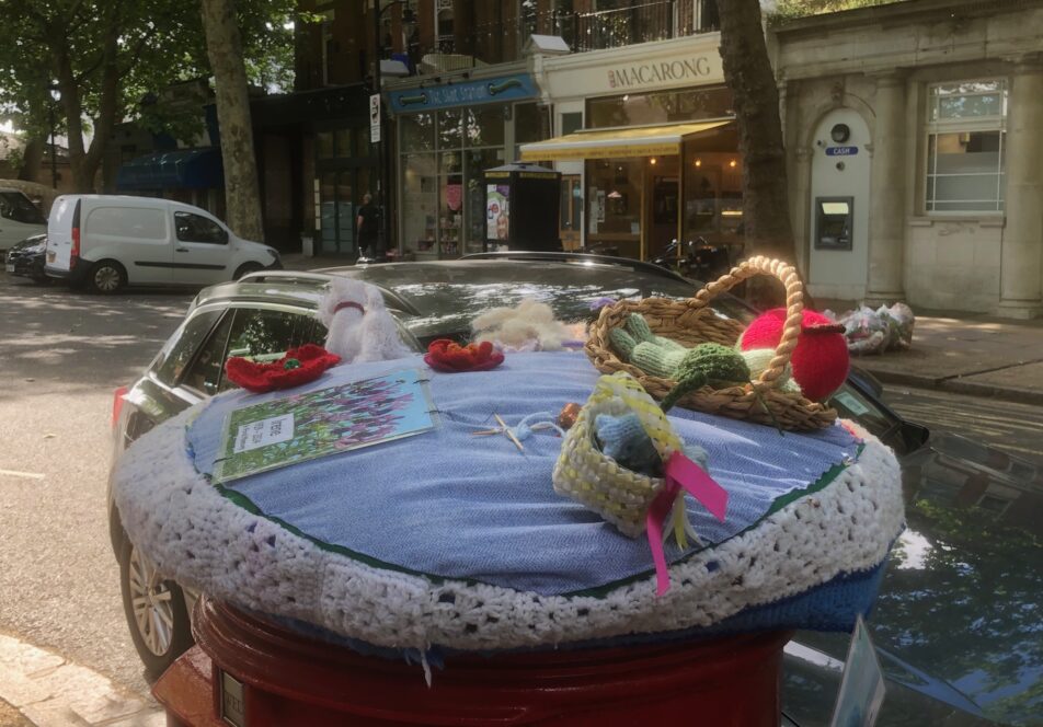 Image shows a knitted topper on a red postbox. The topper includes a knitted basket of fruit, dog, flowers and basket or wool. There is a row of shops in the background and a parked car directly behind the post box.
