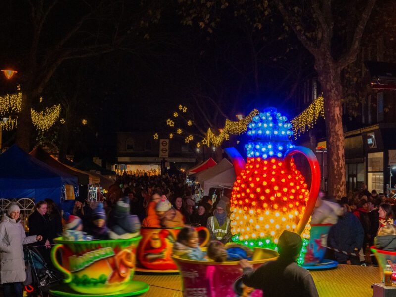 Image from Kew Sparkle showing a brightly lit child's merry go round in the foreground and a street filled with people looking at market stall behind