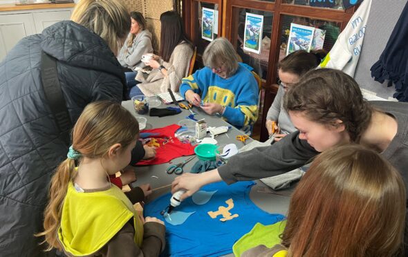 Image shows 6 adults and children around a table crafting. They are working on t-shirts. There is glue, scissors, pieces of fabric and pens on the table