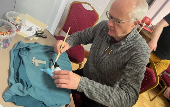 Image shows a white haired balding man wearing a grey jumper. He is seated at a table holding a glue bottle and using a paint brush to apply glue to a t-shirt. There is a t-shirt on the table along with scissors. He appears to be concentrating hard