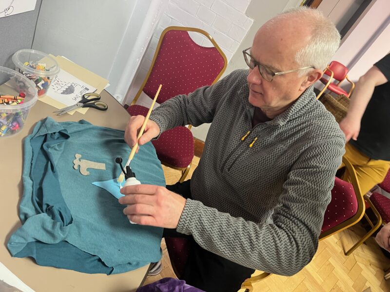 Image shows a white haired balding man wearing a grey jumper. He is seated at a table holding a glue bottle and using a paint brush to apply glue to a t-shirt. There is a t-shirt on the table along with scissors. He appears to be concentrating hard