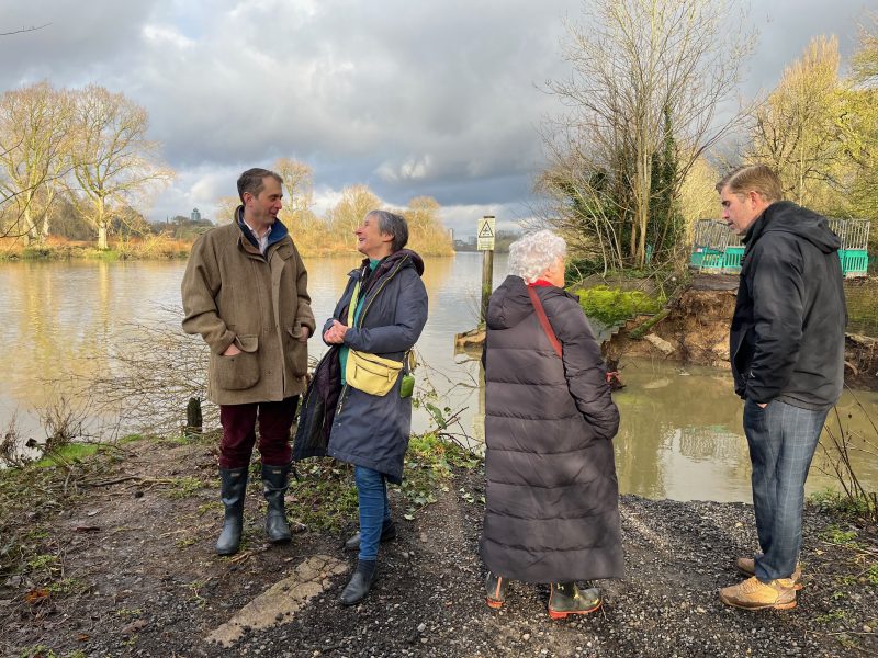 Image shows 4 people in conversation in front of the breach in the towpath. A man and white haired women are in convesation to the right of the image. Another man and women are in conversation to the left of the image. They are all wearing winter coats and walking boots. The sky is grey but the sun is shining. We can see the river in full flood and trees without leaves in the back ground