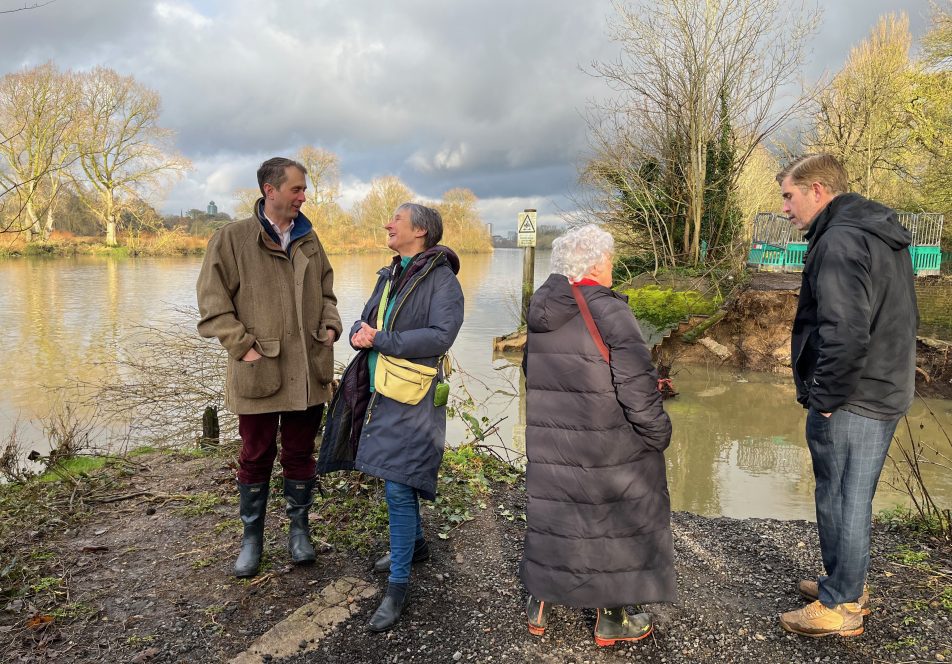 Image shows 4 people in conversation in front of the breach in the towpath. A man and white haired women are in convesation to the right of the image. Another man and women are in conversation to the left of the image. They are all wearing winter coats and walking boots. The sky is grey but the sun is shining. We can see the river in full flood and trees without leaves in the back ground