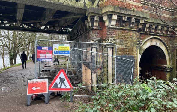 Image shows arch of railway bridge surrounded by metal safety barriers. There is a workman red and white triangle and a pedestrians this way arrow as well as danger keep out signs
