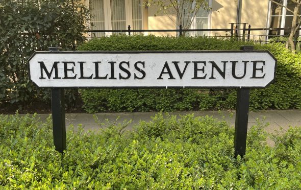 Most of he image is a black and white street sign with the words Melliss Agenue. The sign is standing above a green hedge, in front of a pathway. We can see houses behind