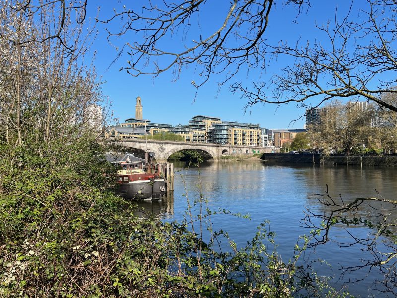 View of two arches of a stone bridge over the river thames. There are blocks of flats behind the bridge and a barge in the foreground along with green bushes. Bare tree branches are set against a bright blue sky