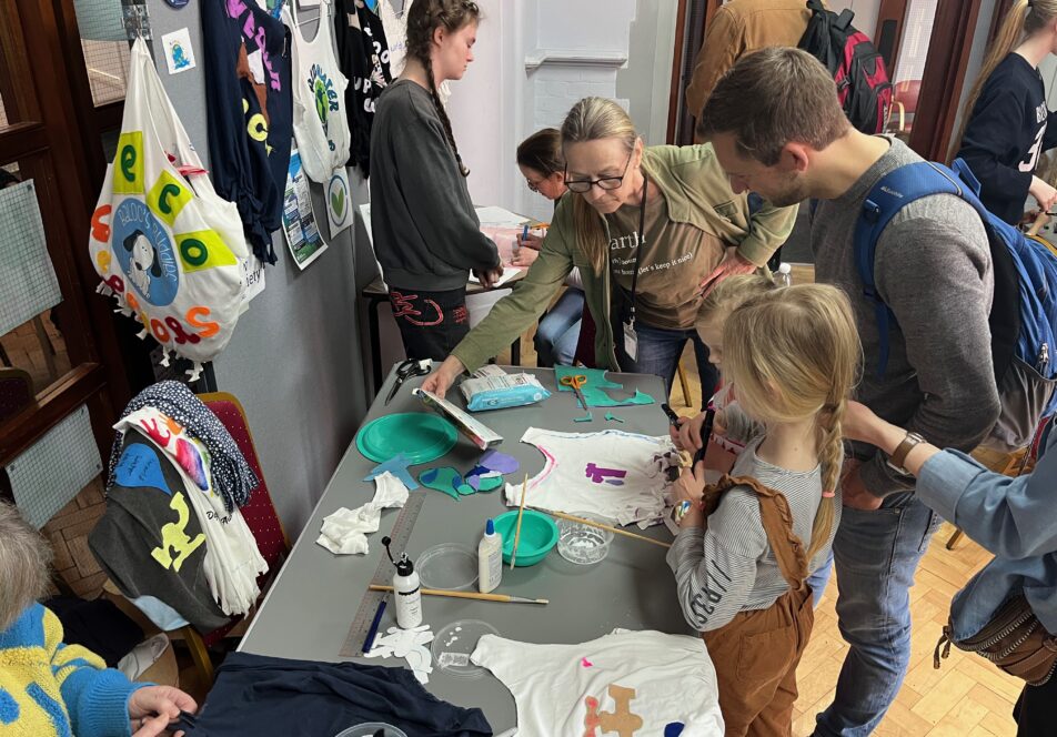 Image shows a busy arts and crafts event with children and adults bending over a table discussing how to make a bag. There cut up t shirts on the table and t shirt bags hung on a board to the left.