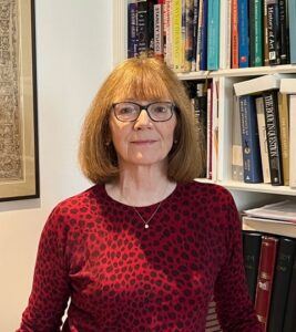 Images shows a middle-aged woman with shoulder length straight red hair looking directly at the camera. We can see her from the waist up. She is wearing a long sleeved deep red and burgundy scooped necked top. It looks like a leopard skin design. She is smiling. She has black rimmed glasses. There is a book case behind her.
