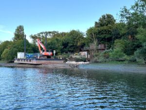 Image shows a large orange digger on a pontoon against a river bank. There are sand bags to the right of the pontoon and evidence of pile driving work beyond that. The river flows in the foreground and there are trees in the background and a clear blue sky