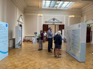 Image shows a large formal looking room with high ceilings and a light brown wooden floor. There are white and blue displays around the room with people standing looking at the words on the displays
