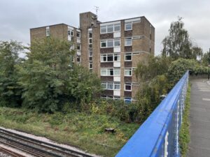 Image shows a 6 storey brown brick block of flats. The windows are white and the cladding underneath them brown. In front of the flats is scrub land with trees, bushes and ivy running up to railway tracks which we can see along the bottom of the picture. There is a blue road bridge cutting across the right.