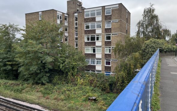 Image shows a 6 storey brown brick block of flats. The windows are white and the cladding underneath them brown. In front of the flats is scrub land with trees, bushes and ivy running up to railway tracks which we can see along the bottom of the picture. There is a blue road bridge cutting across the right.